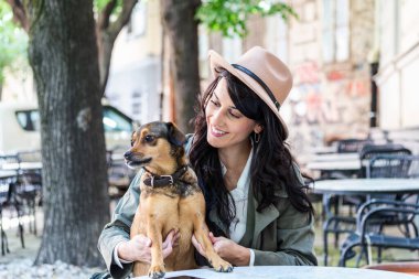Çekici genç bir kadın kahve dükkanında oturuyor, rahatlıyor ve köpeğini tutuyor. Hayvan dostu kafeterya, köpeğiyle bir kafede oturup kahve içen güzel bir kız..