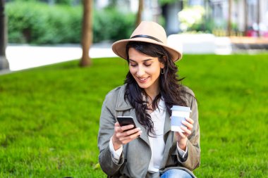 Cep telefonu ve açık havada kahve içen güzel bir kadın. Dışarıda palto giyen, elinde kahve fincanı tutan, cep telefonu kullanan neşeli genç bir kadın.