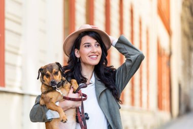 Young smiling woman with a hat in casual clothes iron and hugging dog whilewalking on the street. Smiling young woman enjoying good day and posing with pet. Woman playing with her dog