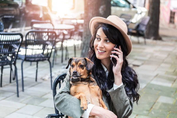 Attractive young woman sitting in coffee shop, relaxing and holding her dog while talking on the phone. Pet friendly cafeteria, beautiful girl with her dog siting at a coffee shop and drink coffee.