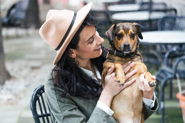 Attractive young woman sitting in coffee shop, relaxing and holding her dog. Pet friendly cafeteria, beautiful girl with her dog siting at a coffee shop and drink coffee.