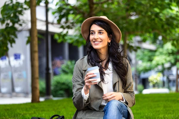Beautiful young woman with cup of coffee outdoors. Cheerful young woman wearing coat outdoors, holding takeaway coffee cup, using mobile phone