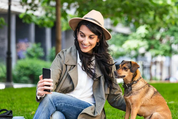 Portrait of pleased girl embracing funny dog and taking a selfie with her mobile phone. Smiling young woman in white shirt enjoying good day and posing with pet