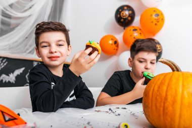 Korkunç kostümlü şirin çocuklar cadılar bayramı keklerinin tadını çıkarıyorlar. Masada balkabağı ve diğer korkunç süslemeler var.