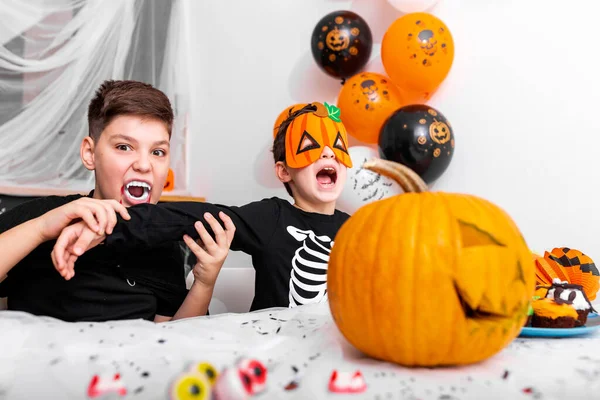 Scary boy with vampire fangs l pretending to bite his brothers arm at the halloween party. Jack O\' Lantern Halloween pumpkin on the table