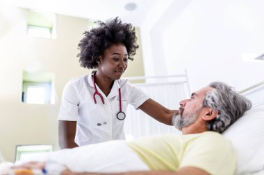 Yatakta yatan kıdemli hasta hastane odasında Afro-Amerikalı kadın doktorla konuşuyor. Sağlık ve sigorta konsepti. Doktor yaşlı hastayı hastane yatağında rahatlatıyor ya da sağlık danışmanlığı yapıyor..