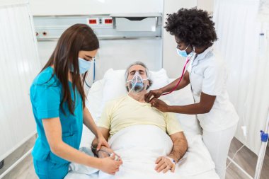 Koruyucu maskeli doktorlar hastanedeki enfekte olmuş yaşlı hastayı muayene ediyorlar. Hastanedeki kıdemli hasta oksijen maskesiyle yatağında dinleniyor..