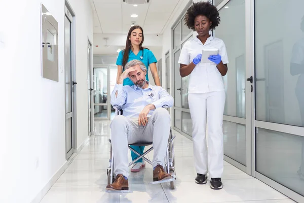Senior man patient in wheelchair sitting in hospital corridor with African American female doctor and nurse. African American doctor and nurse in scrubs with patient in hospital wheelchair in corridor of specialist care clinic