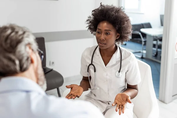 Black female doctor and senior man communicating in a waiting room at hospital. medicine, healthcare and people concept - doctor and patient meeting at hospital