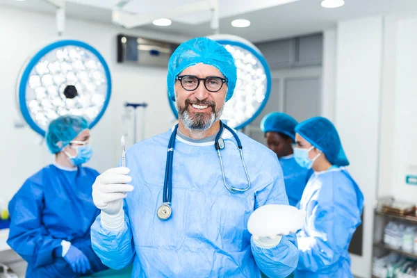 Mature Plastic surgeon man holding silicon breast implants in surgery room interior. Cosmetic surgery concept