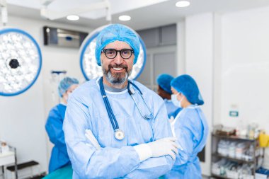 Hastanedeki ameliyathanede kolları çapraz duran erkek cerrahın portresi. Takım cerrahları ameliyat yapıyor, orta yaşlı doktor kameraya bakıyor, modern bir ameliyathanede.