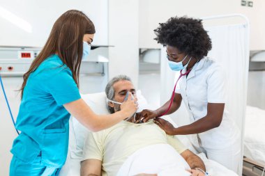 Hemşire hastane yatağında yatan yaşlı bir hastaya oksijen maskesi takarken Afro-Amerikalı doktor da stetoskop, koronavirüs kovidi 19 koruma kavramı ile hastayı muayene ediyor.