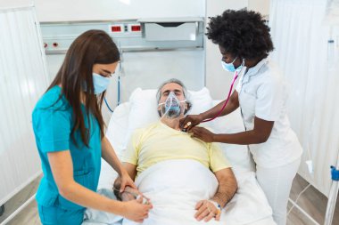 Cerrahi maske takan genç bir doktor ve hemşire Covid-19 salgını sırasında hastane yatağında nefes almasına yardımcı olmak için pozitif basınçlı oksijen maskesi takan yaşlı bir hastayı kontrol ediyor.