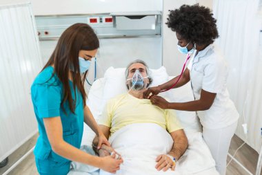 Koruyucu maskeli doktorlar hastanedeki enfekte olmuş yaşlı hastayı muayene ediyorlar. Hastanedeki kıdemli hasta oksijen maskesiyle yatağında dinleniyor..