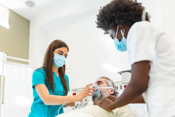 Nurse puts oxygen mask on elderly man patient lying in the hospital room bed while African American doctor examining patient with stethoscope, coronavirus covid 19 protection concept