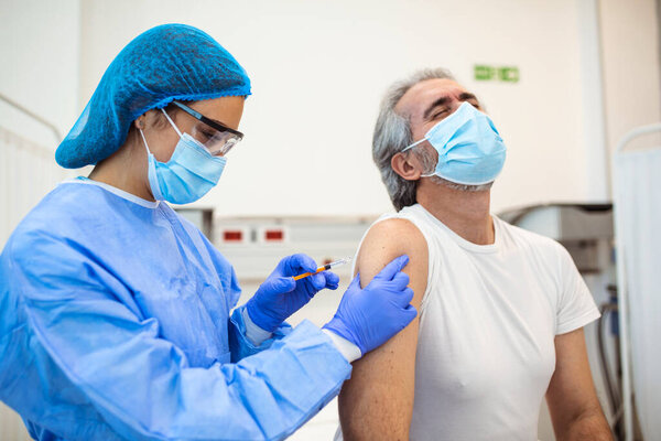 Doctor in gloves holding syringe and making injection to senior patient in medical mask. Covid-19 or coronavirus vaccine. Doctor vaccinating male patient in clinic, closeup