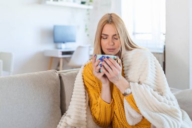 beautiful stylish woman sitting at home thinking, romantic melancholy mood, wearing knitted sweater, feeling warm, winter style, drinking coffee, covered in woolen blanket