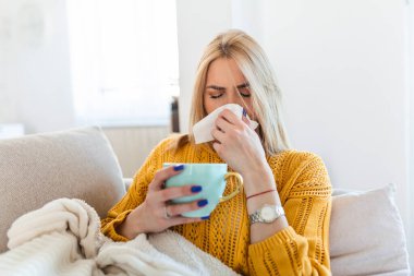 Hasta kadın. Flu. kadın üşüttü. Dokuların içine hapşırmak. Baş ağrısı. Virüs. İlaçlar. Burnunu mendile sümküren soğuk algınlığına yakalanmış genç bir kadın. Kanepede oturan, başı ağrıyan hasta kadın.