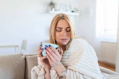 Close up of dreaming young woman sitting in living room with cup of coffee or tea enjoying under blanket
