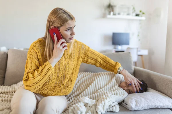 Worried mother taking temperature of her little son who is lying in bed with fever. Young mother checking the temperature of her boy with thermometer on a couch in the living room , calling doctor.