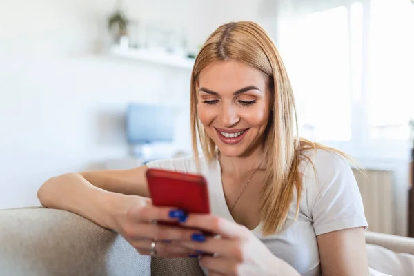oung beautiful woman sits on the couch with a phone in her hand texting. Young woman sit relax on sofa in living room browsing surfing wireless internet on smartphone,