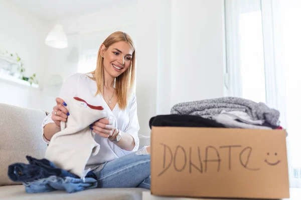 Young woman with donation box at home. Donation box for poor with clothing in female hands. Woman donates clothing to shelter. Cheerful woman holds box of donated clothing