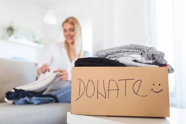 Woman packing clothes into donation box in living room. Girl puts in a box with donations items. Volunteering. Woman participating at charity and holding donation box