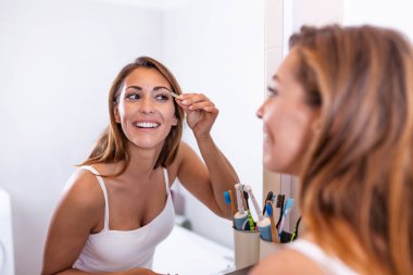 Kişisel bakım ve makyaj konsepti. Banyo aynasında aynadan önce beyaz gömlekli genç bir kadının kaşlarını cımbızla yolarken belini kaldır.