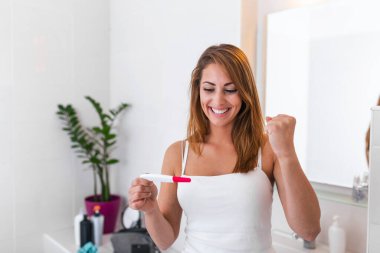 Beyaz bluz giymiş, hamilelik testinin sonucuna bakıp gülümseyen ve bebek sahibi olacağı için mutlu genç bir bayanın yakın çekimi. Sonunda hamile..