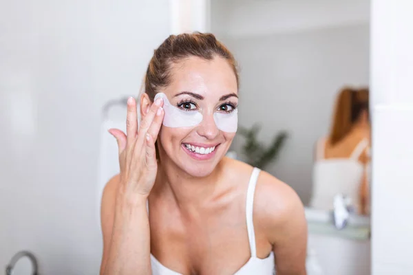 Beauty woman applying under-eye mask looking herself in the mirror in the bathroom. Skin care girl touch patches of fabric mask under eyes to reduce eye bags.