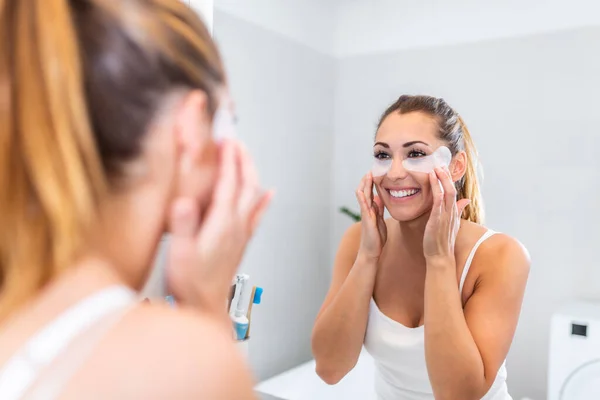 young woman after shower looking in mirror pampering herself applying face mask use anti-aging beauty product feeling happy, skin care, everyday morning routine concept