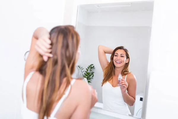Self care concept. Waist up reflection of young smiling woman in white shirt enjoying her morning while applying favorite perfume before bathroom mirror
