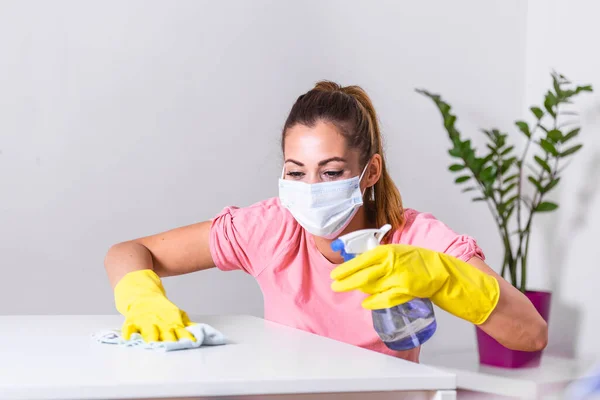 Woman with protective glove and facial mask doing housework, housewife portrait. She sprinkling disinfectant and cleaning the table. Stay safe.