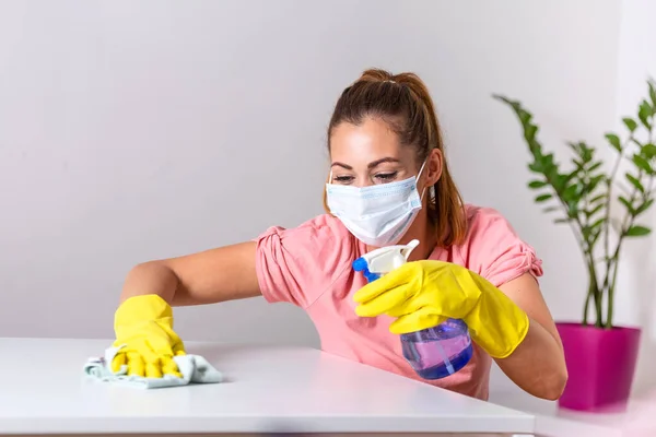 Woman with protective glove and facial mask doing housework, housewife portrait. She sprinkling disinfectant and cleaning the table. Stay safe.