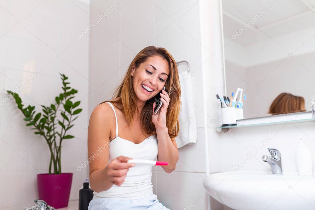 Smiling woman sitting in bathroom and holding positive pregnancy test and talking on her smart phone with the father to give him happy news. Smiling pregnant girl telling good news on mobile.