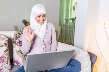 Young Muslim woman having video call via laptop at home. Happy smiling Muslim woman sitting on sofa, couch and using laptop at living room at home, learning language, video calling.