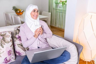 Diz üstü bilgisayarını kullanan Müslüman bir kadın, Evde Sosyal Ağ Sörf, Boş Alan. Evde internette sörf yapmak. Laptopta çalışan ve evinde kanepede otururken gülümseyen çekici Müslüman kadının yan görüntüsü.
