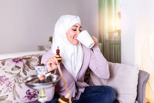 Muslim woman smoking shisha at home and drinking coffee or tea. Muslim young woman enjoying while smoking nargile. Arab girl smoking Hookah