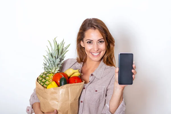 Young woman holding paper bag full of groceries and showing her mobile phone, Order online concept. shopping app