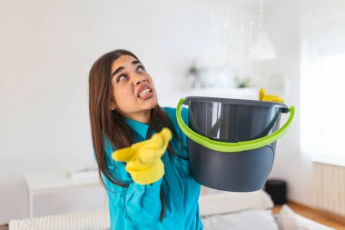 Shocked Woman Looks at the Ceiling While Collecting Water Which Leaks in the Living Room at Home. Worried Woman Holding Bucket While Water Droplets Leak From Ceiling in Living Room