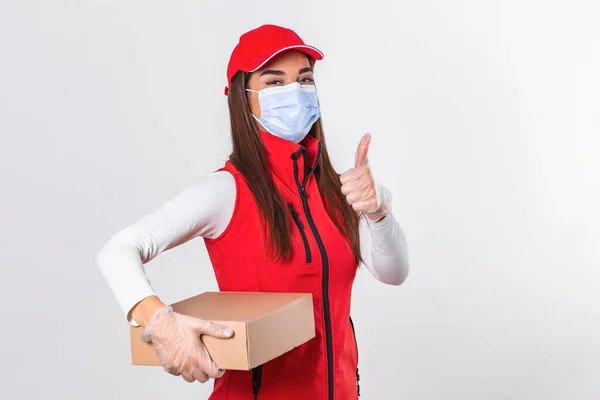 Delivery woman employee in red cap blank t-shirt uniform face mask gloves hold cardboard box showing thumbs up isolated on white background. Quarantine pandemic coronavirus virus 2019-ncov concept