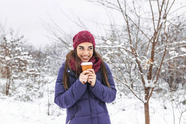 Pretty young woman in winter outfit holding disposable cup filled with hot coffee or tea. Girl holding mug of hot beverage in her hands and walks outdoors in winter.