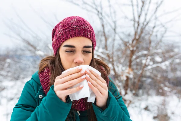 Young woman suffering from a seasonal cold and flu blowing her nose on a handkerchief as she stands outdoors on winter. Healthcare and medical concept