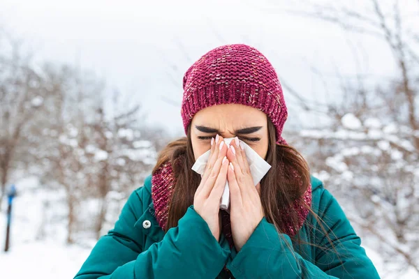 Young woman sick and sneeze into tissue paper. Girl blowing nose outdoors.Catching cold in winter. Upset gloomy woman with ill expression sneezes and has running nose