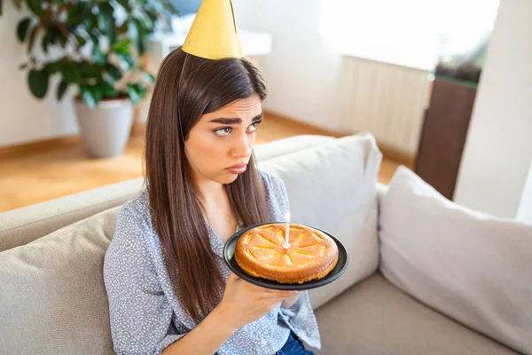 Virtual Party During Lockdown. Upset and frustrated single african american woman in party hat celebrating birthday online. Lady making video conference, holding cake with candle, sitting on the floor