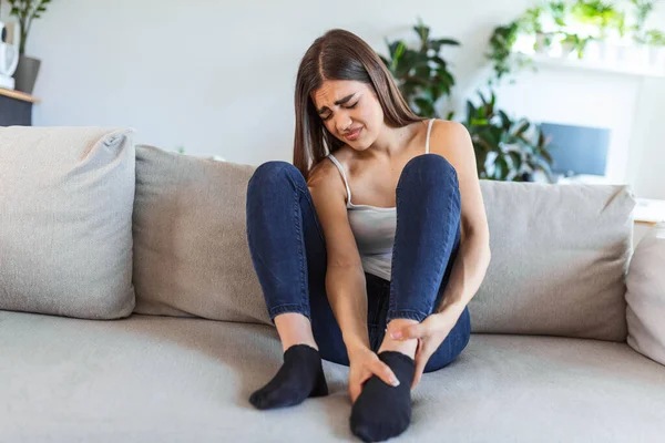 Hands massaging swollen foot while sitting on sofa during the day at home. Photo of Young Caucasian woman suffering from pain in leg. Woman massaging her legs after all day at work in office