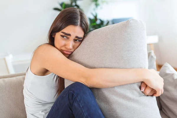 Young woman hugging pillow with depression and crying on sofa at home. Lonely millennial lady feeling stressed and hopeless, suffering from mood disorder or having psychological problem