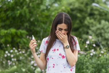 Young pretty woman sneezing in front of blooming tree. Spring allergy concept. Woman applies nasal spray. Pollen allergy symptoms