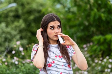 Pollen allergy concept. Young woman using nose spray for her pollen and grass allergies. Flowering trees in background. Spring Seasonal allergies and health problems.