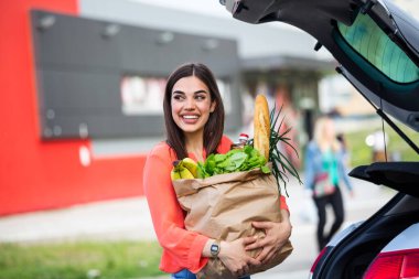 Young woman riding shopping cart full of food on the outdoor parking. Young woman in car park, loading shopping into boot of car. Shopping successfully done. Woman putting bags into car after shopping